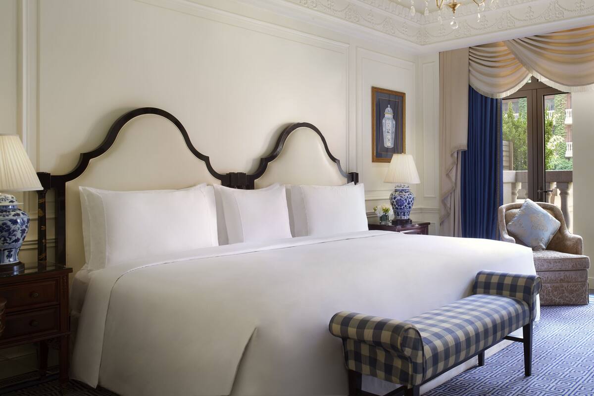A large bed with a dark headboard, two bedside tables with lamps, a bench, and a view of the outdoors through a window.