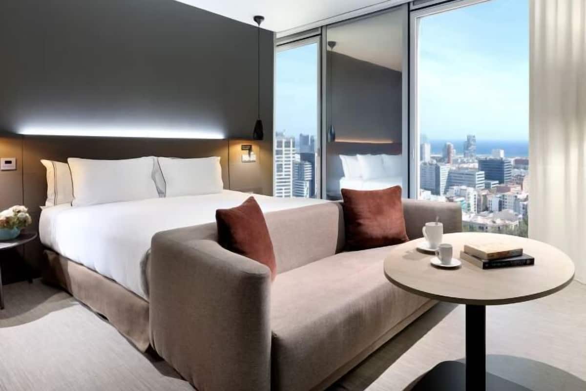 A modern hotel room with a large bed, a sofa, a round table, and a city view.