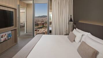 Habitación de hotel con una cama grande, televisión, puerta corrediza a un balcón con vistas a la ciudad y un mueble para revistas.