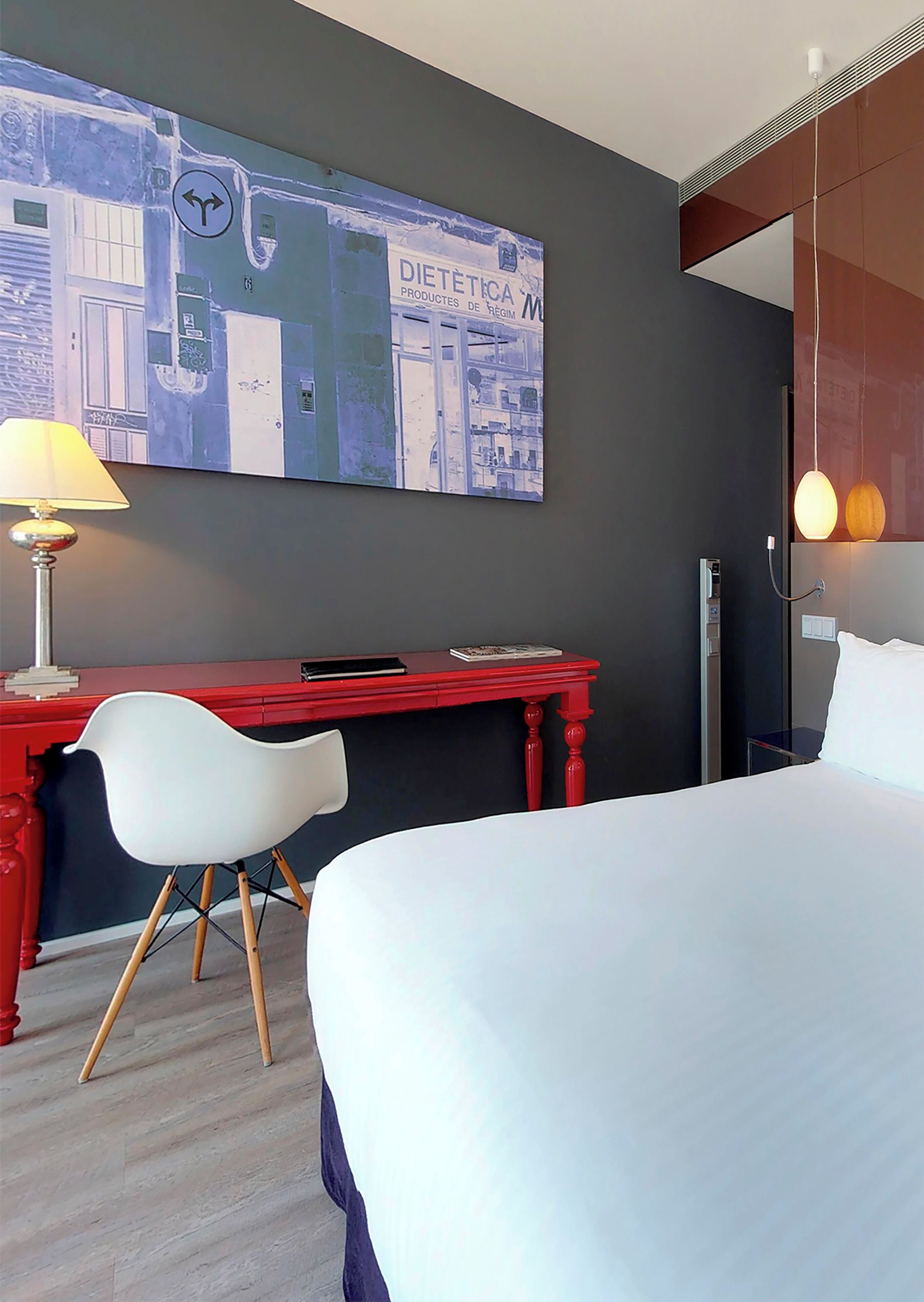 Una habitación de hotel con cama, escritorio, silla, lámpara y una obra de arte en la pared.