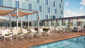 Un área de piscina en la azotea con sillones y sombrillas, una terraza de madera y un edificio moderno con grandes ventanales al fondo.