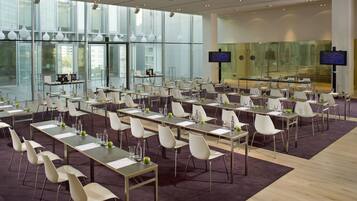 Een congreszaal met meerdere tafels en stoelen, grote ramen en twee schermen aan de muur.