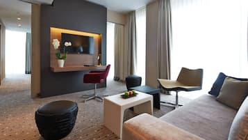 A modern hotel room with a large flat-screen TV mounted on a wooden wall, a comfortable sofa, a red chair, a small table with fruit, and a large window with curtains.