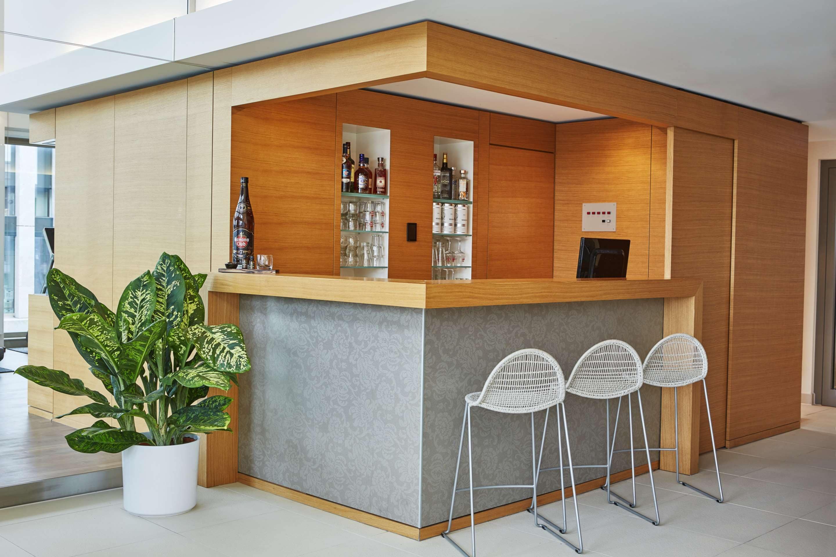 A modern reception area with a wooden counter, glass shelves with bottles, and three white wire chairs.