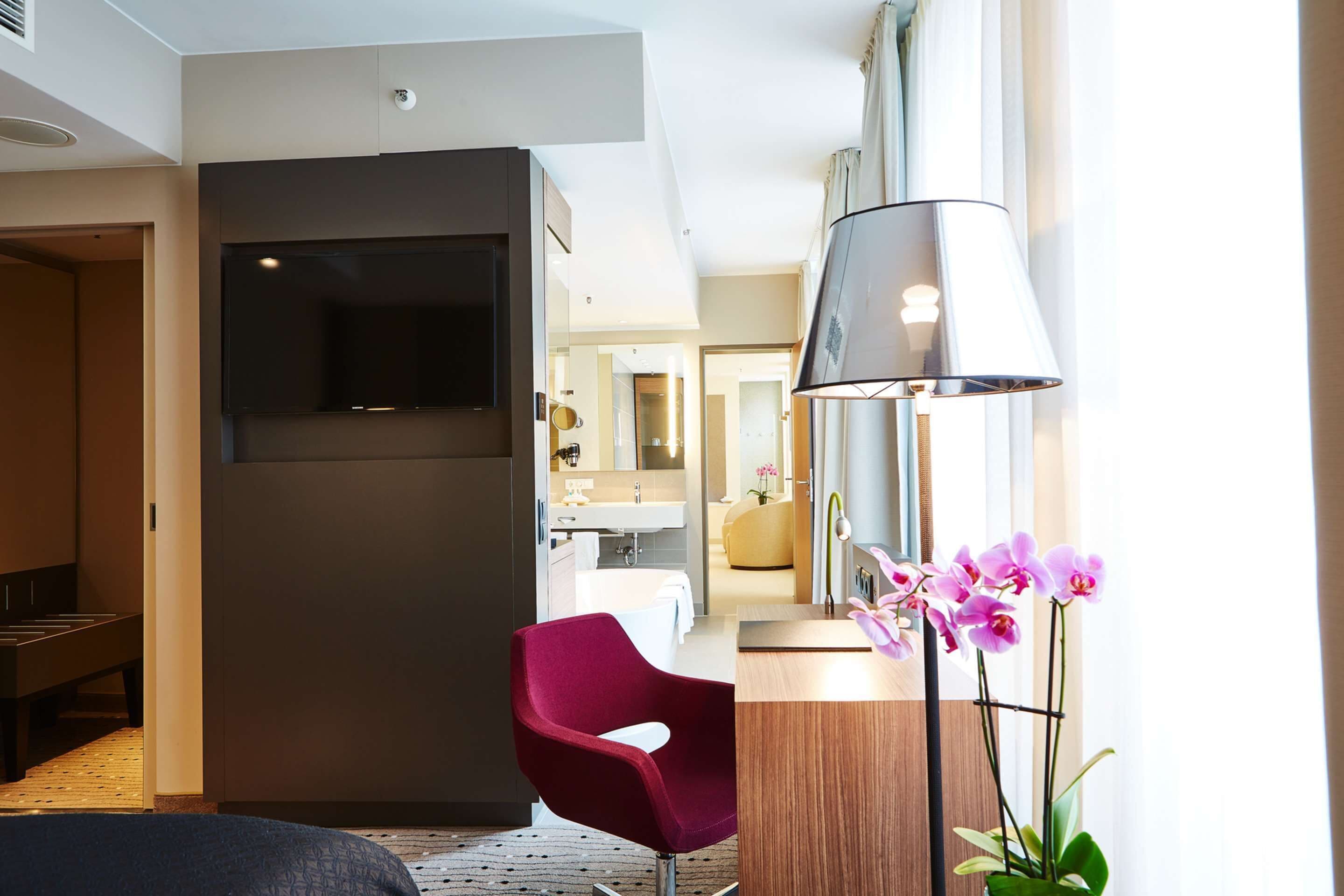 Habitación de hotel moderna con un televisor de pantalla plana grande, una silla roja, una mesita de noche de madera y una orquídea en maceta.
