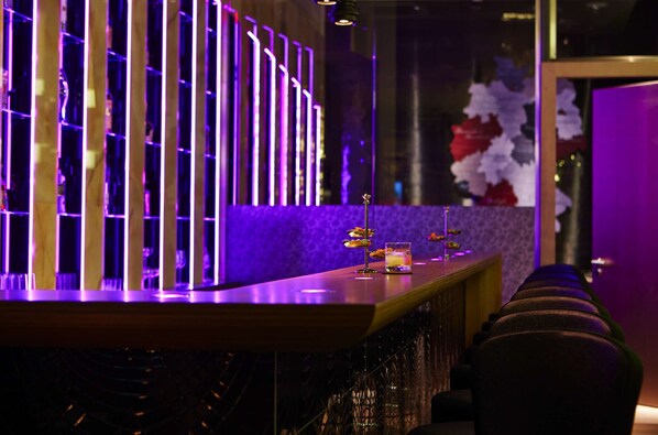 A bar area with a long counter, illuminated by vertical neon lights, and a decorative wall with a floral arrangement.