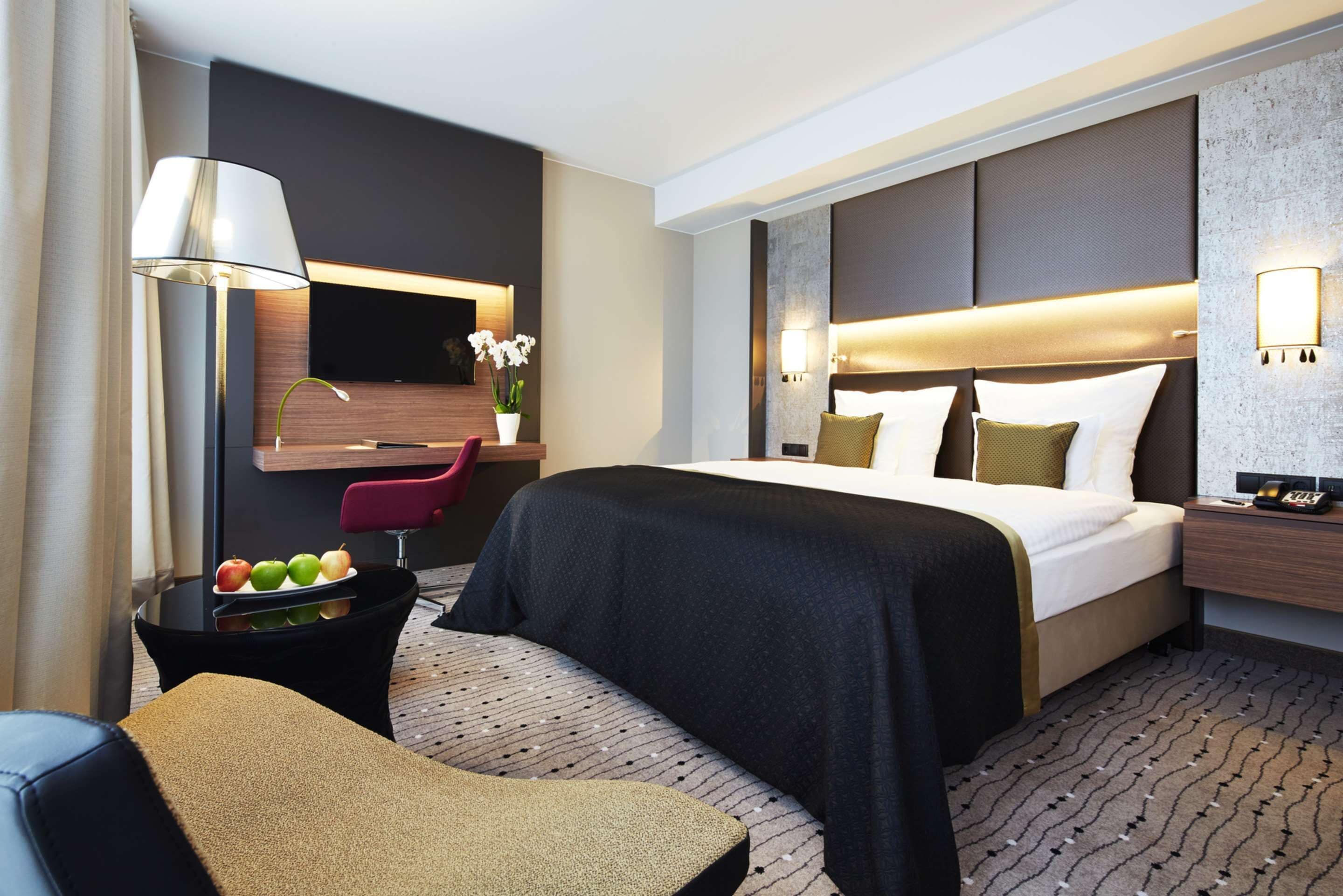Une chambre d’hôtel moderne dotée d’un grand lit, d’un bureau avec un téléviseur à écran plat, d’une chaise et d’une petite table sur laquelle se trouve des fruits.