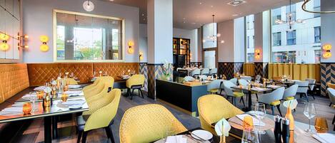 A modern restaurant with high ceilings, pendant lighting, and a mix of yellow and grey upholstered chairs.