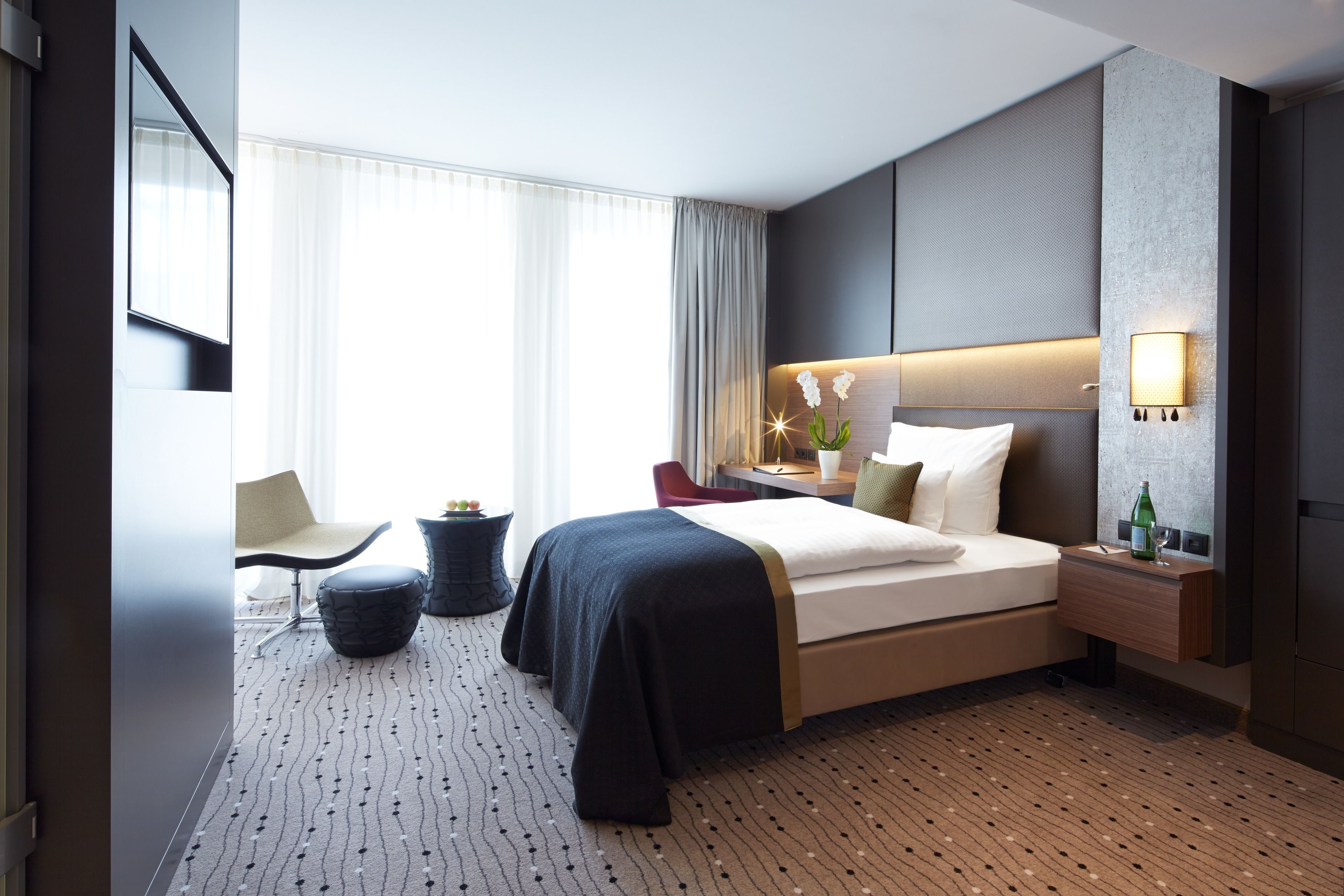 Una habitación de hotel moderna con una cama grande, un escritorio, una silla y una mesita de noche con lámpara.