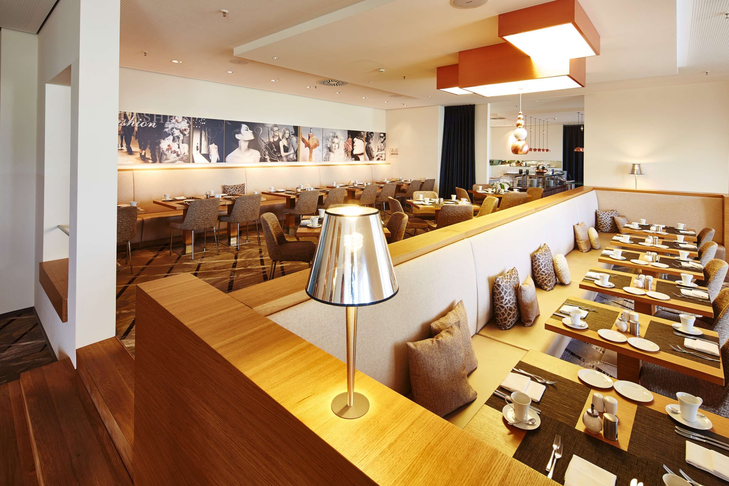 A modern hotel dining area with multiple tables and chairs, a central bar area, and a large wall with photographs.