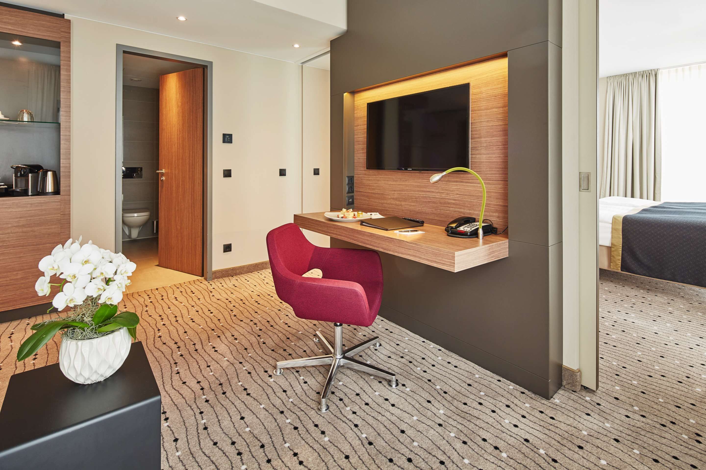 A modern hotel room with a wooden desk, a red chair, a large mirror, and a potted orchid.