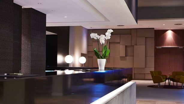 A modern reception area with a dark reception desk, a white vase with white flowers, and yellow chairs. - Steigenberger Hotel Am Kanzleramt (Berlin)