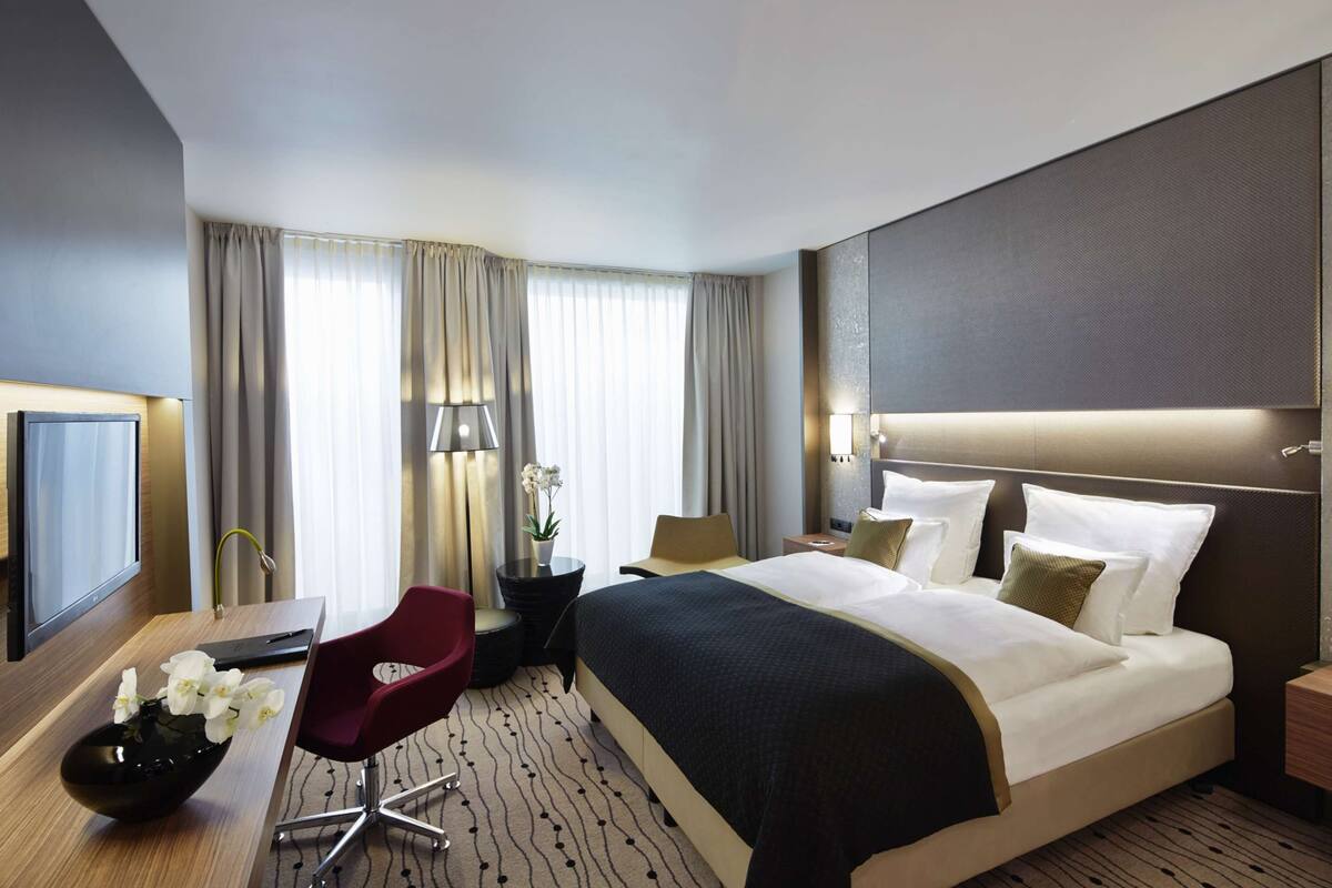 A modern hotel room with a large bed, a desk with a chair, a TV, and a decorative vase.