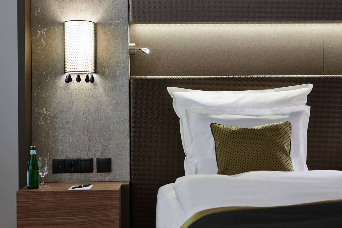A neatly made bed with white and green pillows, a bedside table with a bottle and glass, and a wall-mounted light fixture.