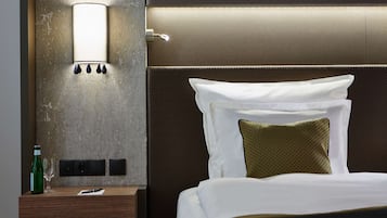 Een net opgemaakt bed met witte en groene kussens, een nachtkastje met een fles en glas, en een wandlamp.