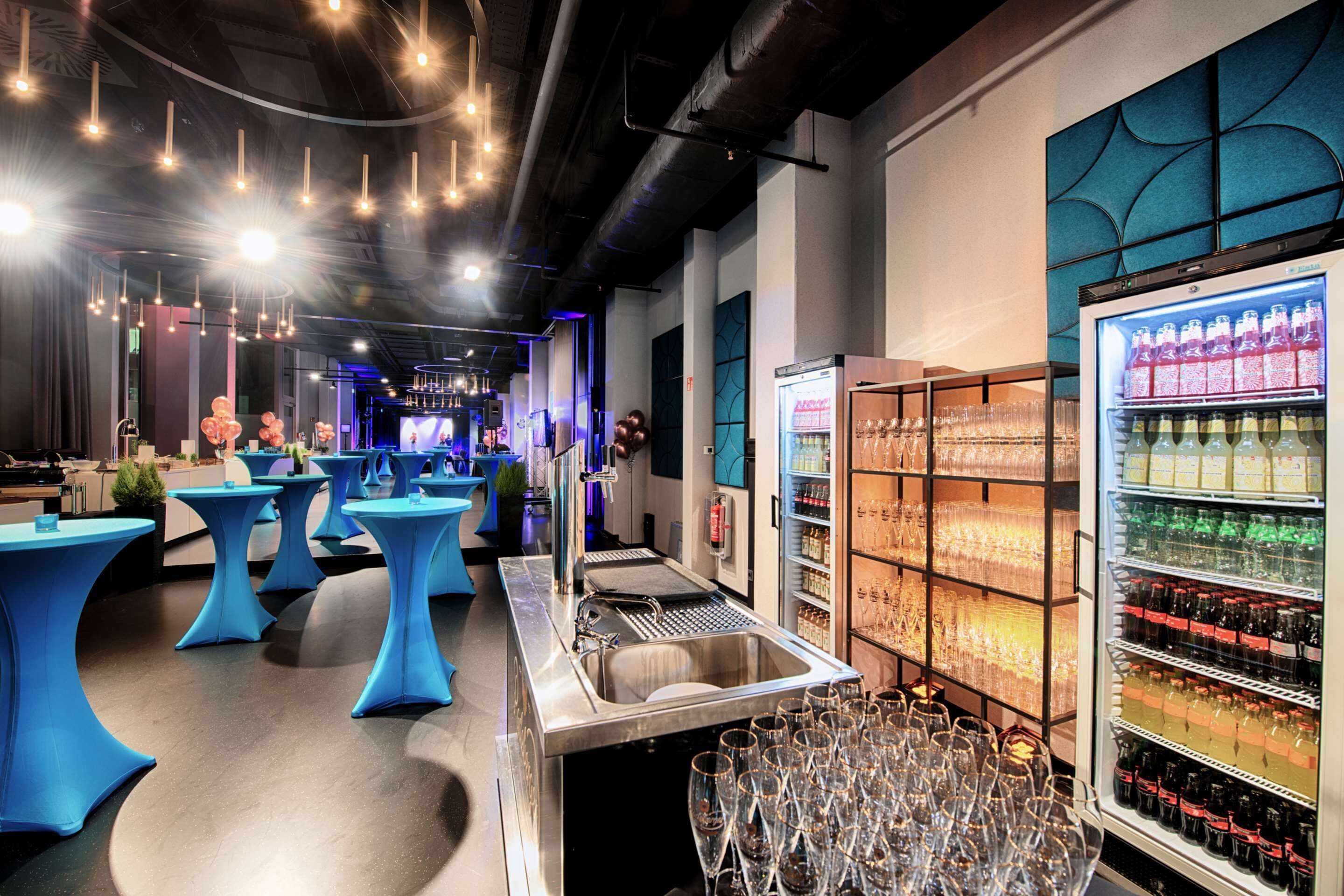 A bar area with blue tables, a bar counter, and a refrigerator stocked with bottles.