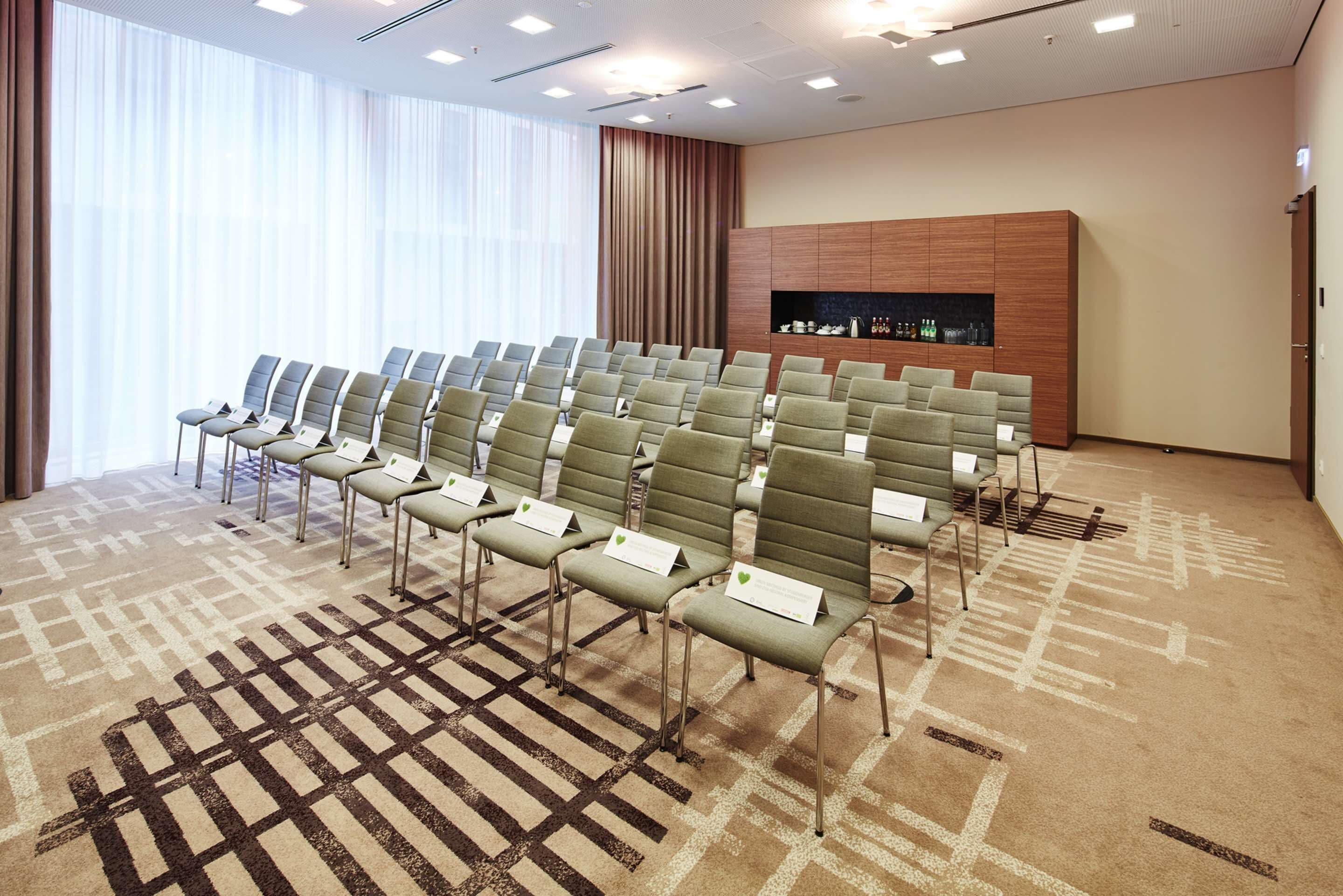 Un salón de conferencias con filas de sillas, piso alfombrado, un mueble de madera con botellas y un ventanal con cortinas.
