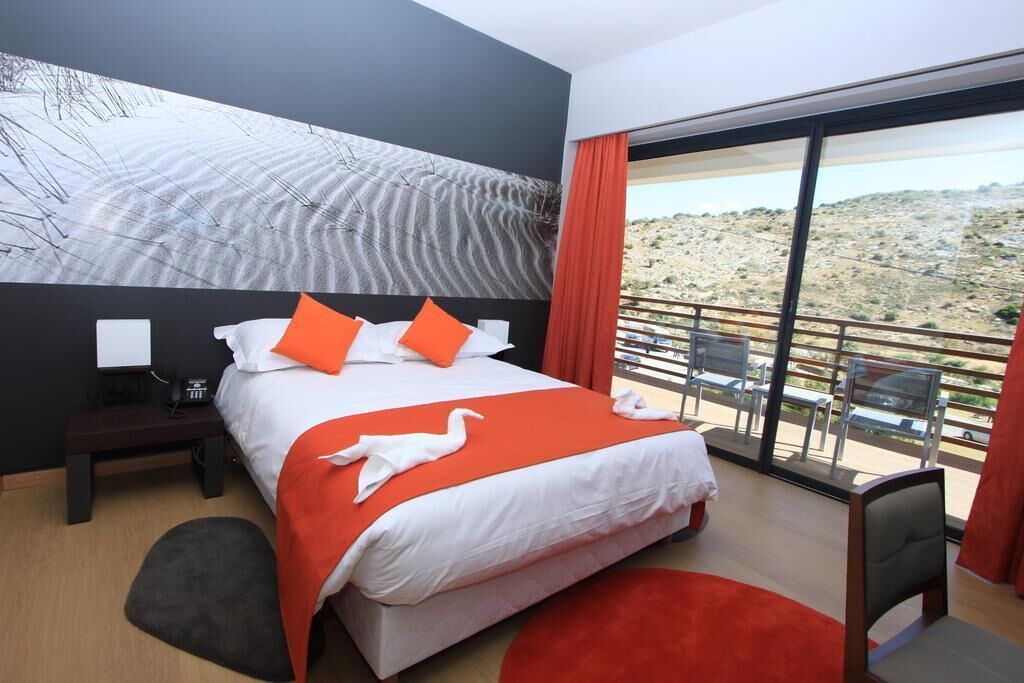 Une chambre d’hôtel avec un grand lit, une table de chevet, un bureau et une vue sur un paysage désertique, visible à travers de grandes fenêtres.