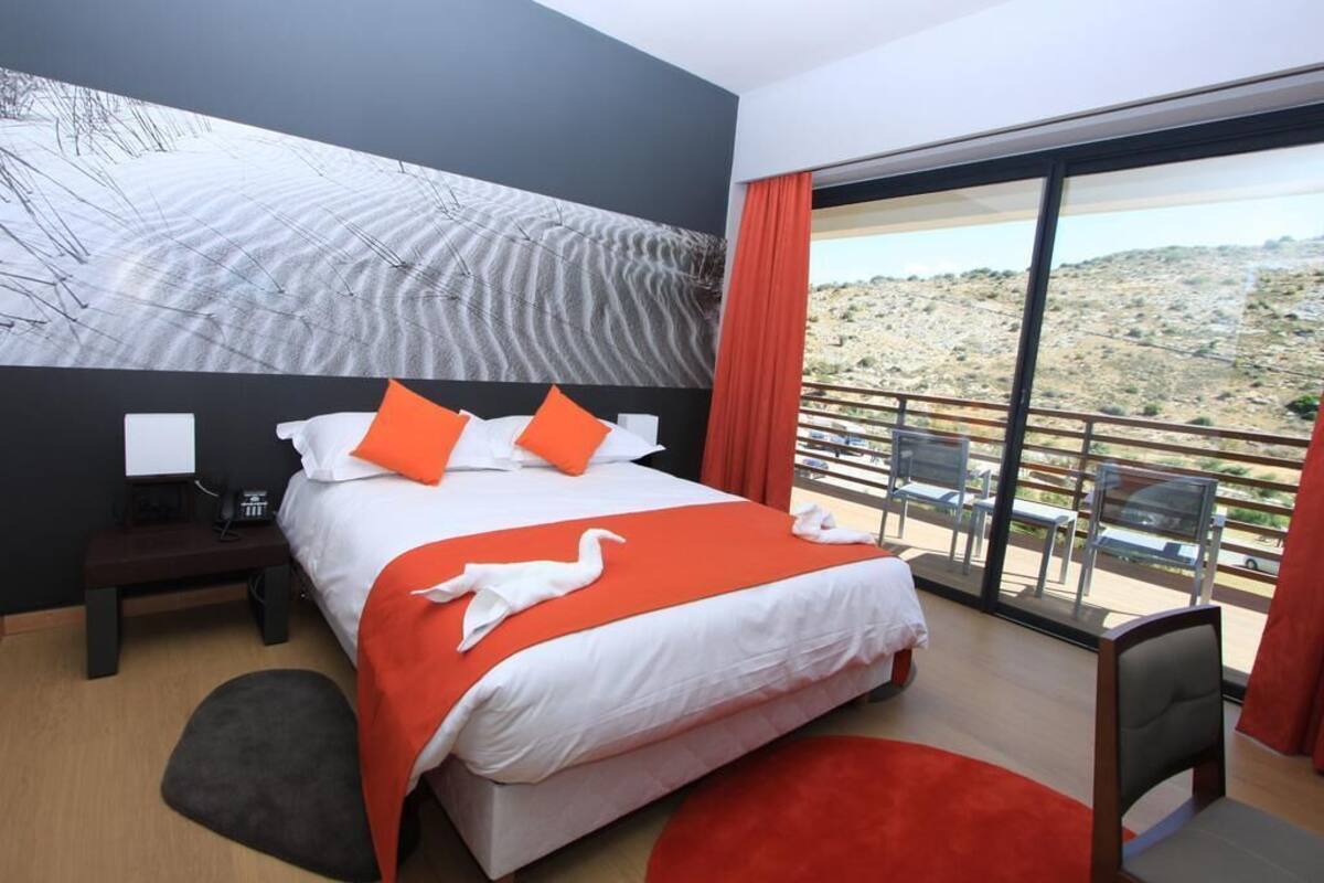 A hotel room with a large bed, a nightstand, a desk, and a view of a desert landscape through large windows.