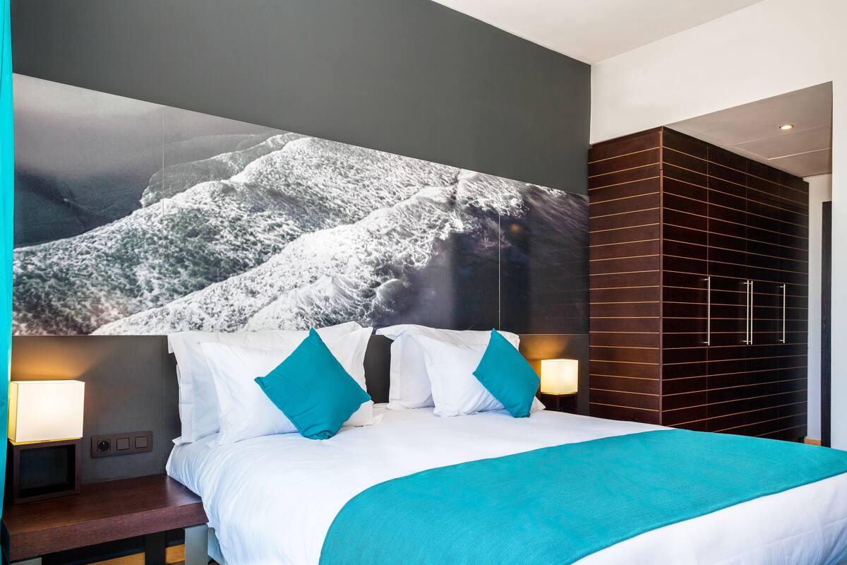 A modern hotel room with a large bed, two nightstands, a wall-mounted TV, and a dark wooden wardrobe.
