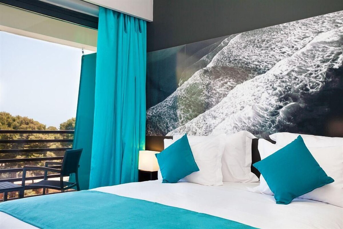 A hotel room with a large bed, teal curtains, and a view of the outdoors.
