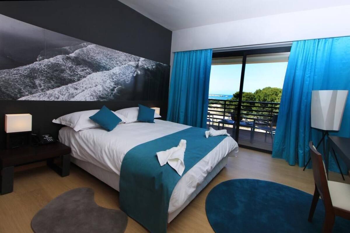 A hotel room with a large bed, a view of the outdoors, and a balcony with blue curtains.