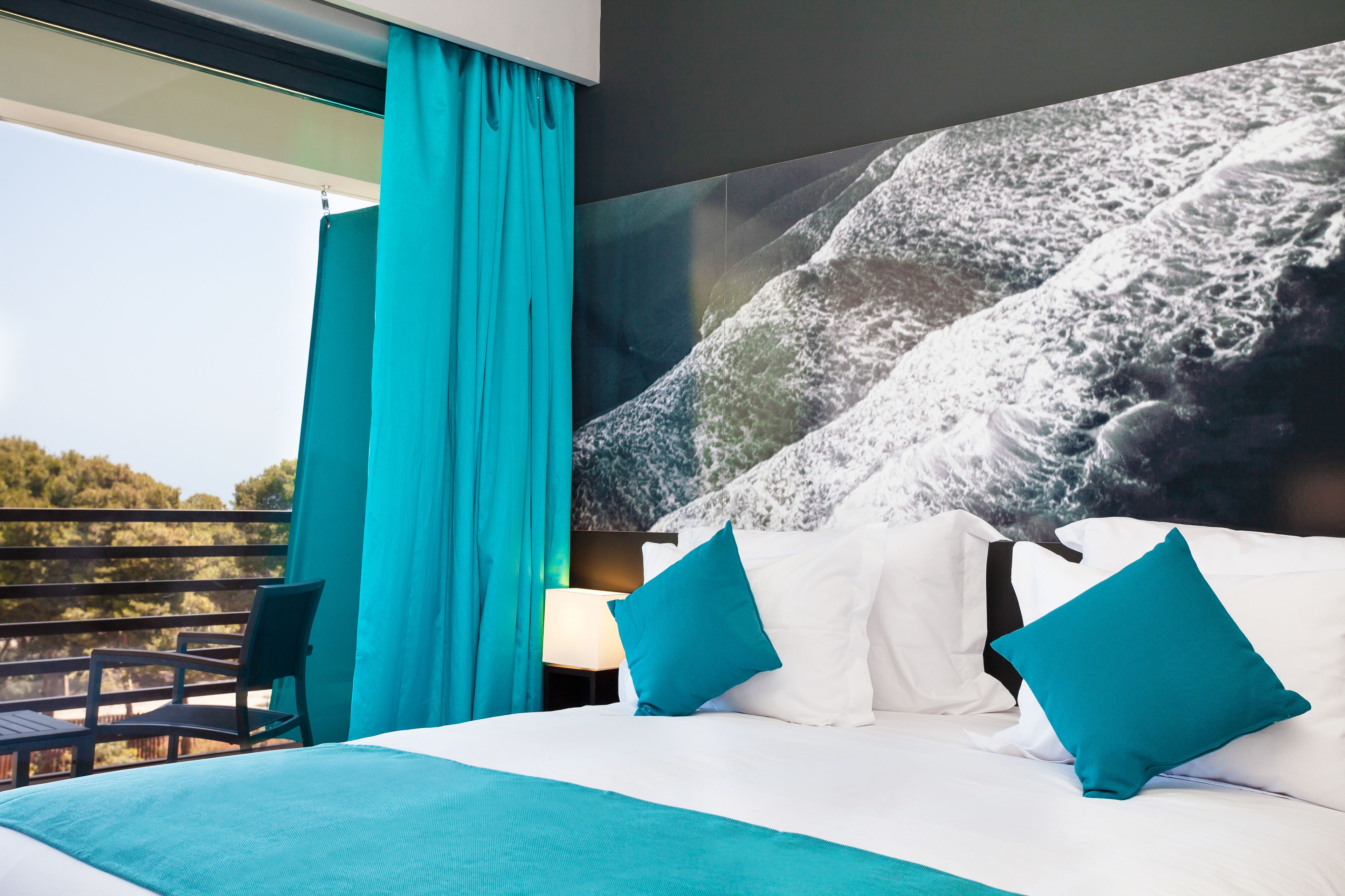 Ein Hotelzimmer mit Bett, blaustichigen Vorhängen, Blick ins Freie und einem großen Poster an der Wand.