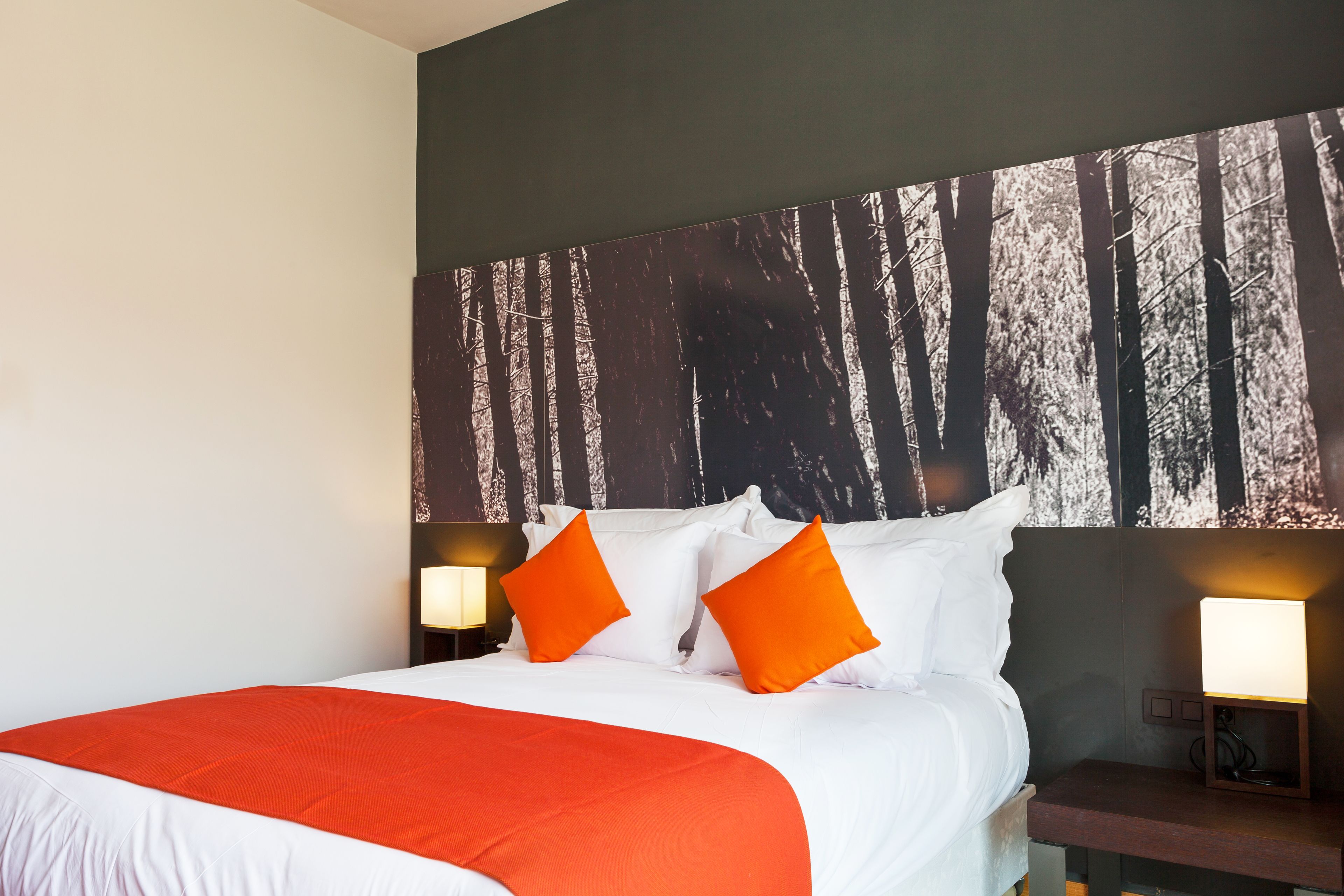 Habitación de hotel con una cama grande, almohadas anaranjadas, edredón blanco y un mural en la pared con motivos forestales.
