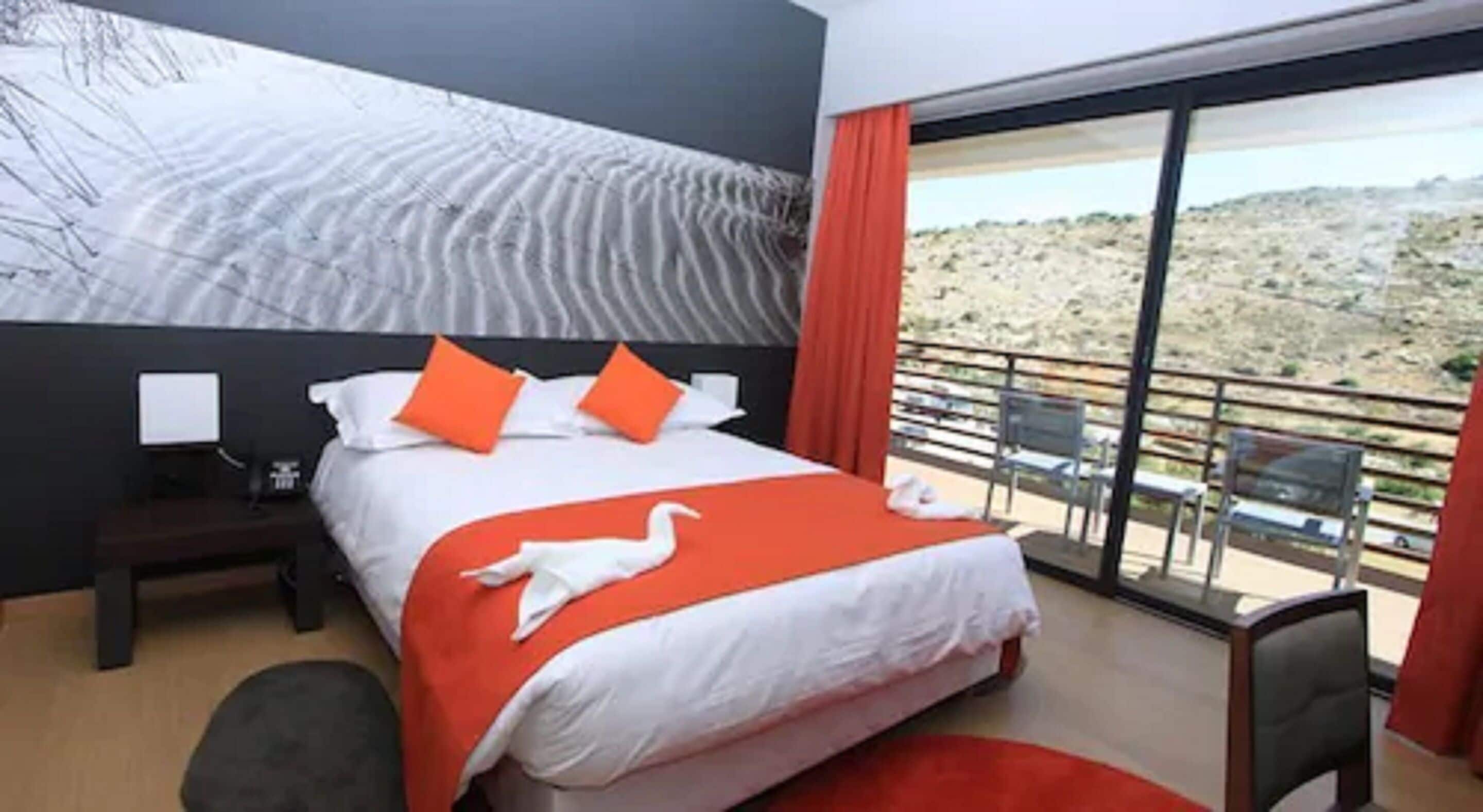 A hotel room with a large bed, a nightstand, a desk with a computer, and a balcony with a view of the landscape.