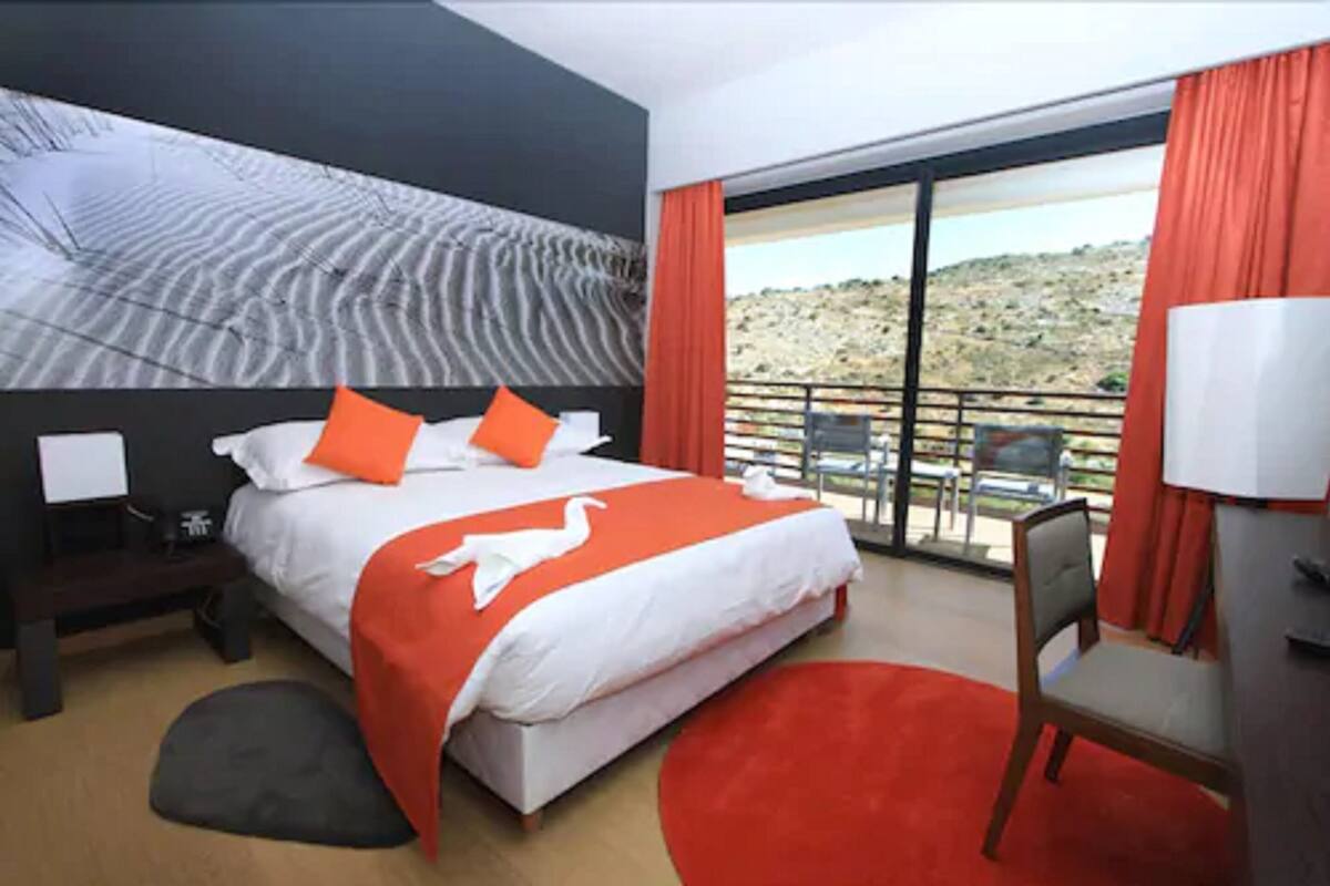A hotel room with a large bed, a desk, and a balcony with a view of the hills.