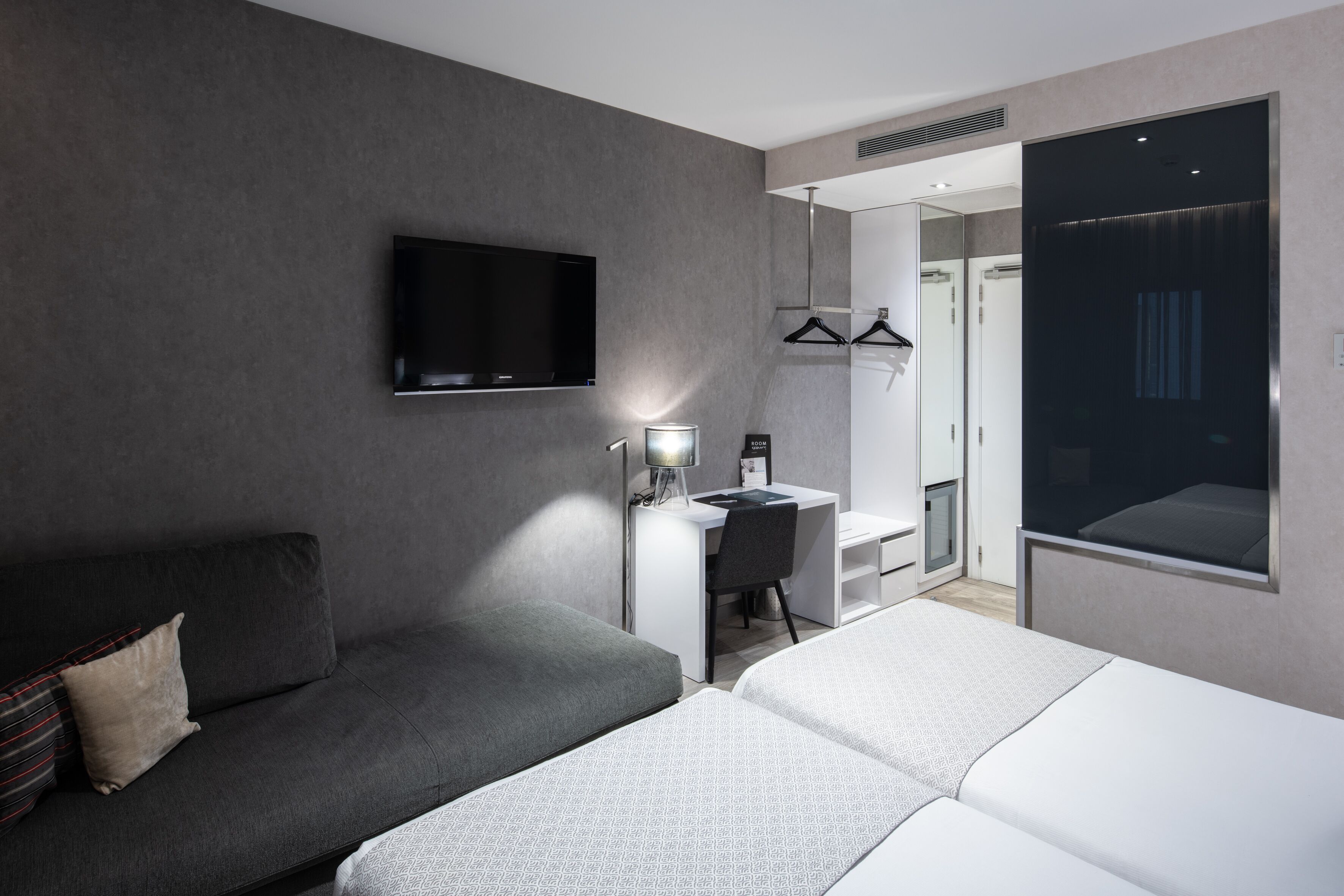 Una moderna camera d'hotel con un divano grigio, un letto, una scrivania con una sedia, una TV fissata al muro e un grande specchio.