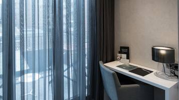 A hotel room with a desk, chair, and a view through sheer curtains.