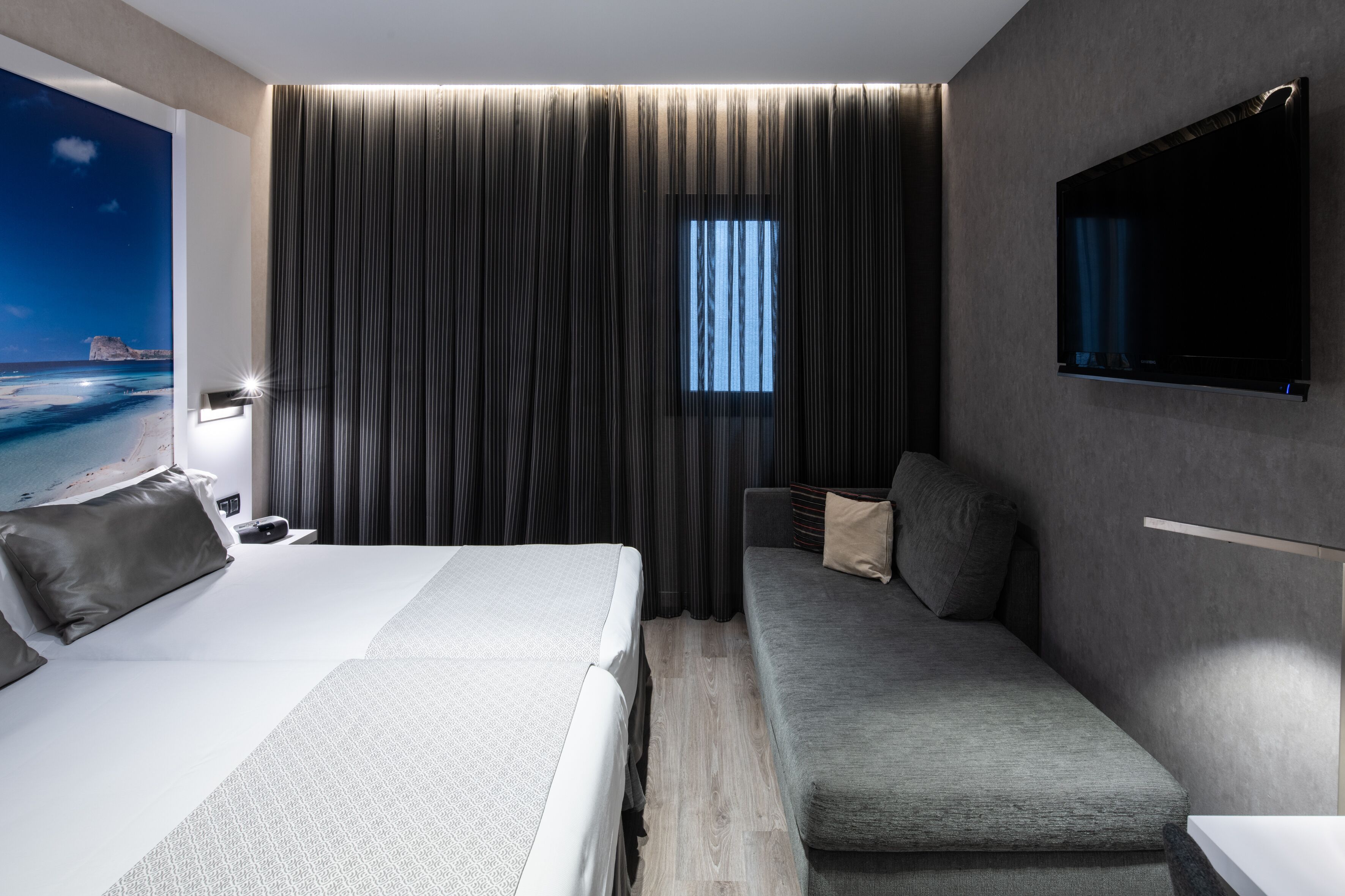 A modern hotel room with a large bed, a sofa, a TV, and a window with curtains.