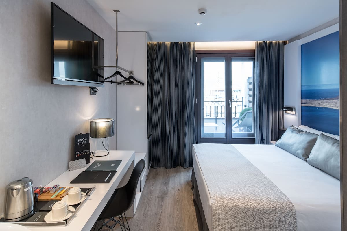 Ein Hotelzimmer mit einem Bett, einem Schreibtisch mit Computer, einem Fernseher und Blick auf die Stadt durch ein großes Fenster.