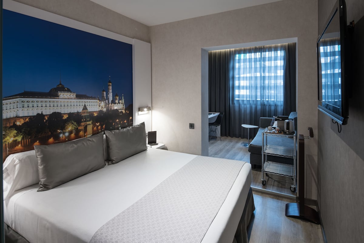 Ein Hotelzimmer mit einem großen Bett, einem Schreibtisch mit Stuhl, einem kleinen Tisch, einem Wagen mit Gegenständen und einem großen Fenster mit Vorhängen.