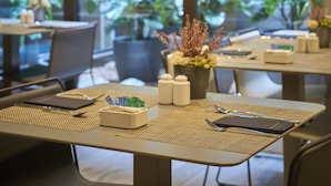 A modern dining area with tables set for two, featuring woven placemats, cutlery, and a notebook. Large windows provide a view of lush greenery outside. - Catalonia Gran Vía BCN (Barcelona)