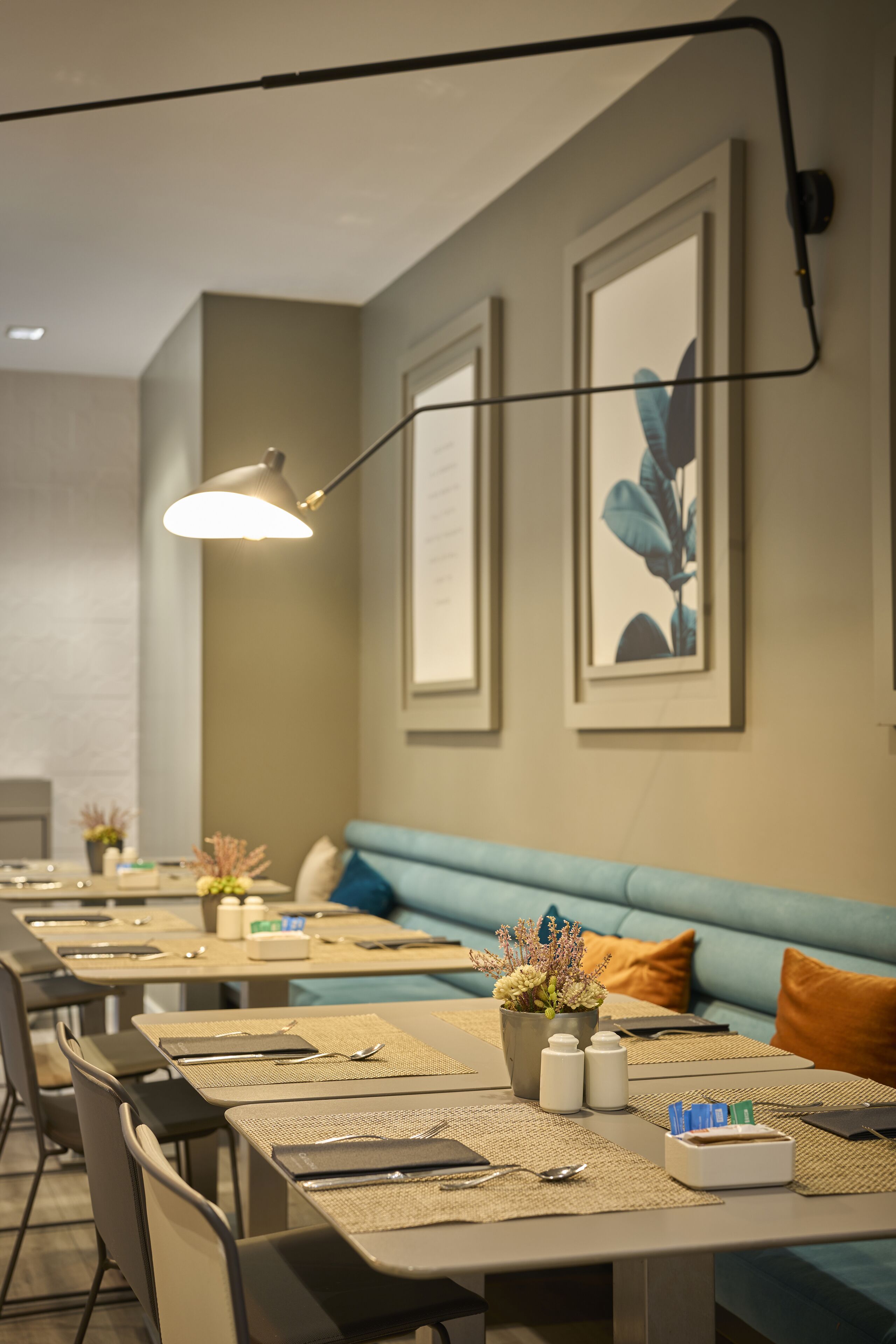 A dining area with tables set for two, each with a placemat, cutlery, and a small vase of flowers. There are cushioned benches along the wall and a hanging light fixture above the tables.