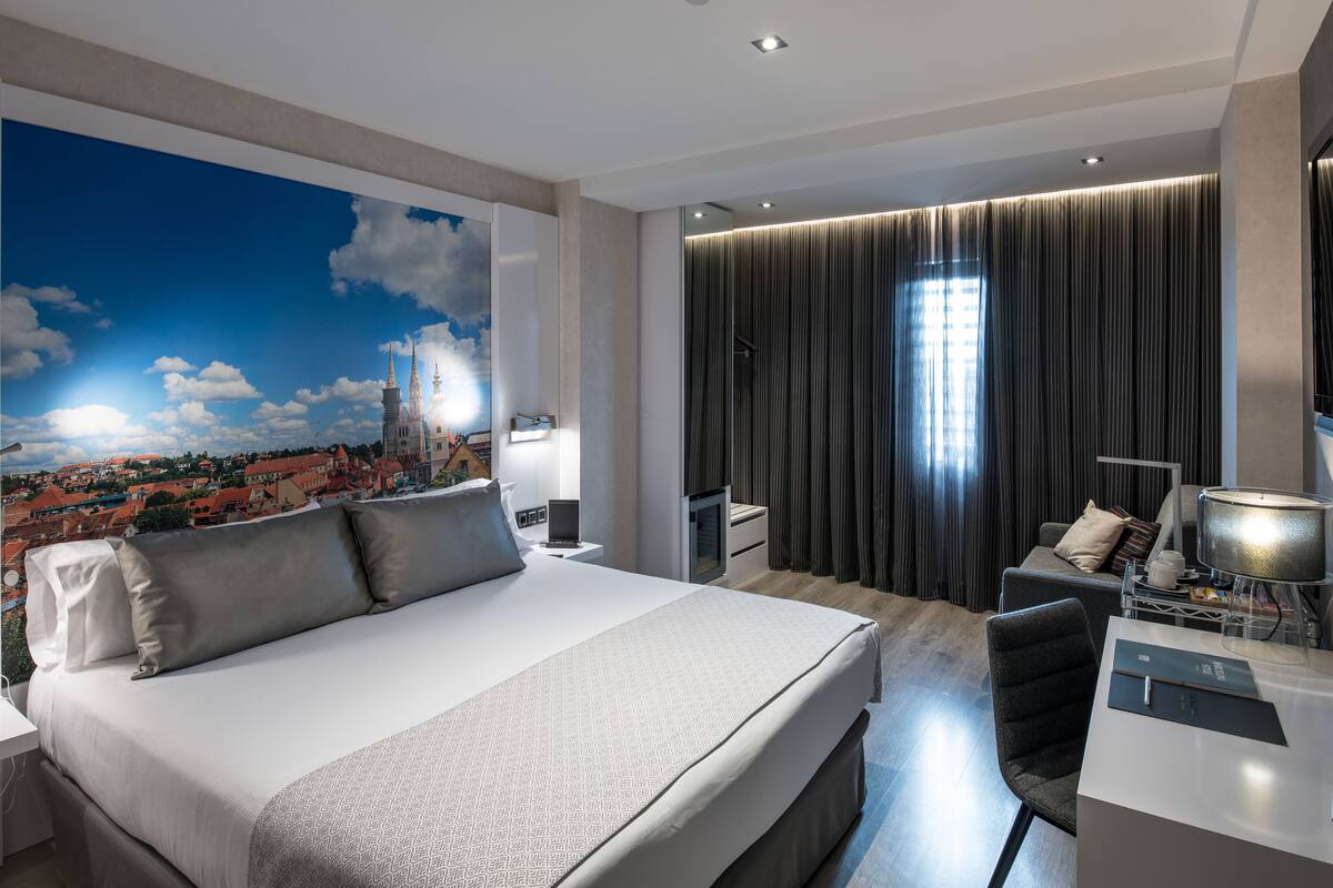 Ein modernes Hotelzimmer mit einem großen Bett, einem Schreibtisch, einem Sessel und einer Stadtansicht als Wandbild.