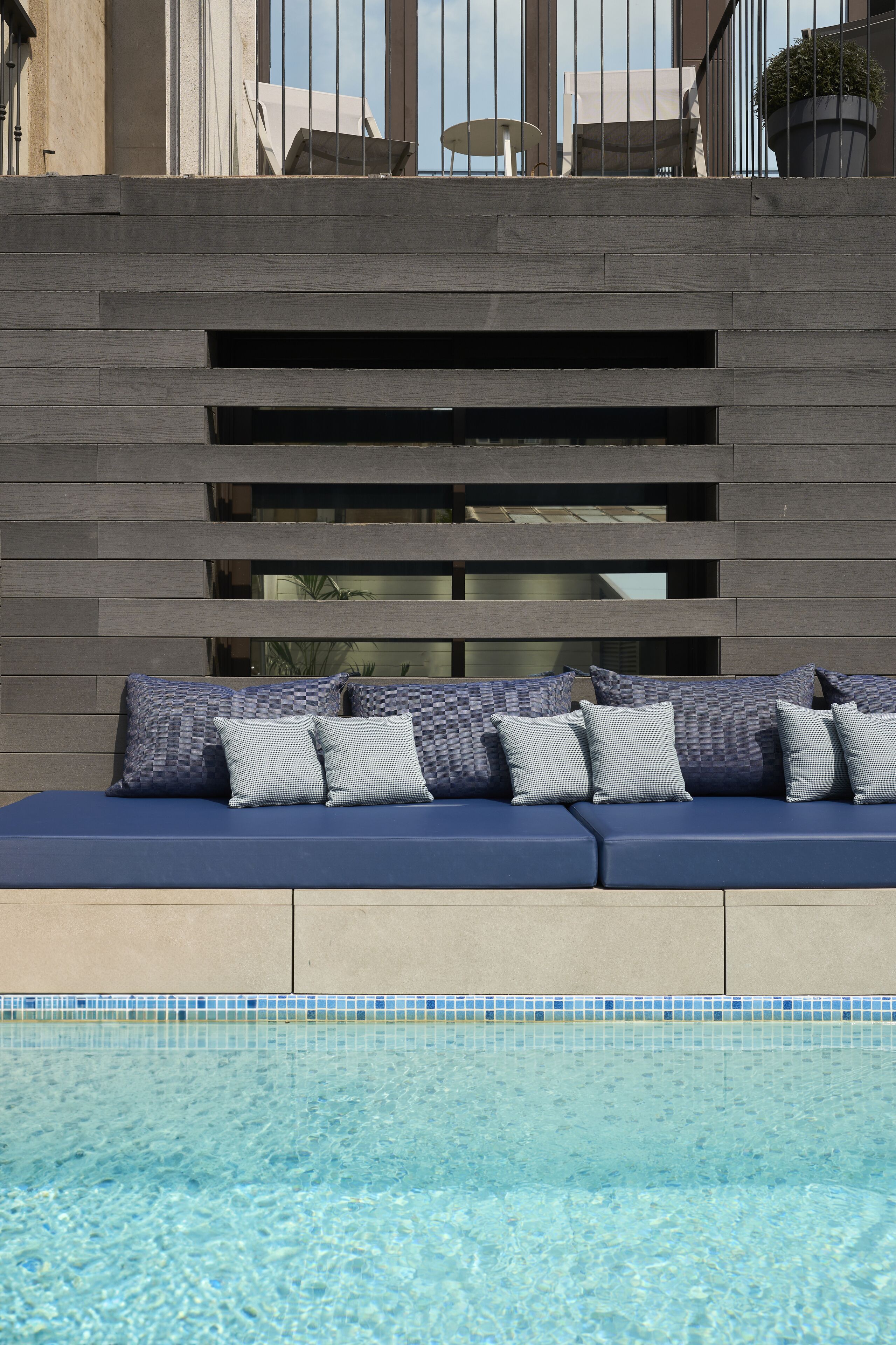A poolside seating area with a built-in bench and cushions, a tiled pool edge, and a modern building facade in the background.
