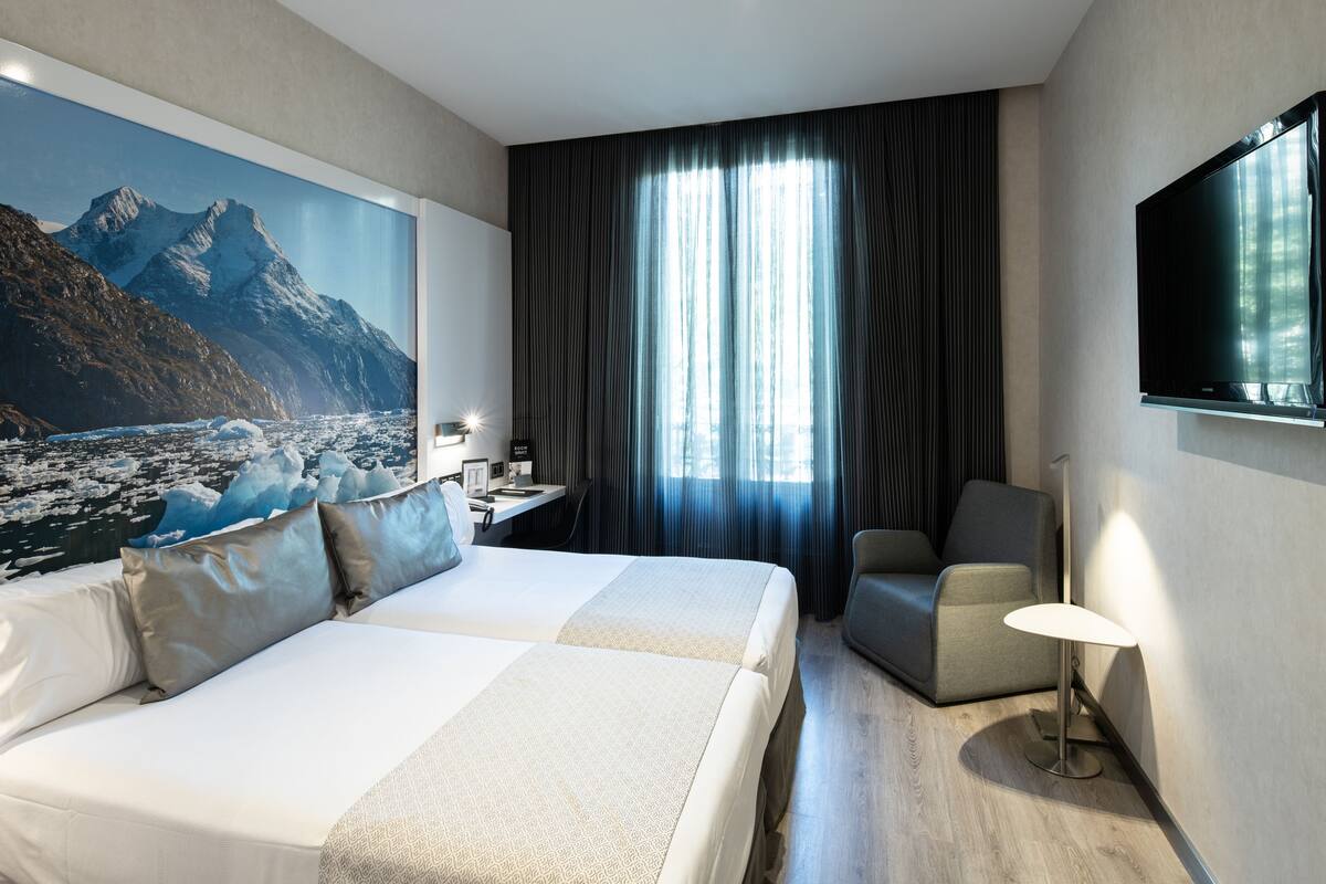 Ein Hotelzimmer mit Bett, Schreibtisch, Sessel, Fernseher und einem großen Fenster mit Vorhängen.