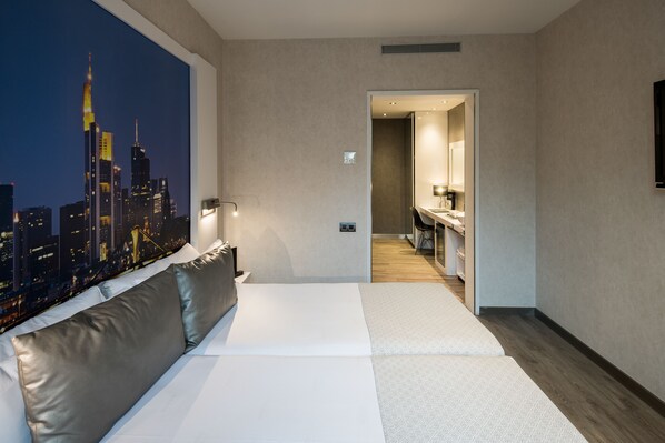 Une chambre d’hôtel moderne avec un grand lit, une fresque représentant un paysage urbain sur le mur, une petite kitchenette et une salle de bain visible à travers une porte ouverte.