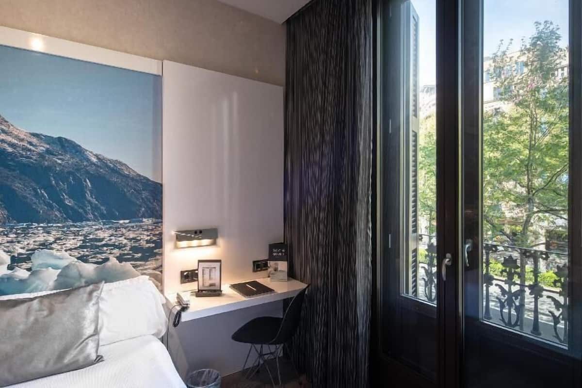 Ein Hotelzimmer mit Bett, Schreibtisch mit Computer, Fenster mit Vorhängen und einer Wand mit einem Bergbild.