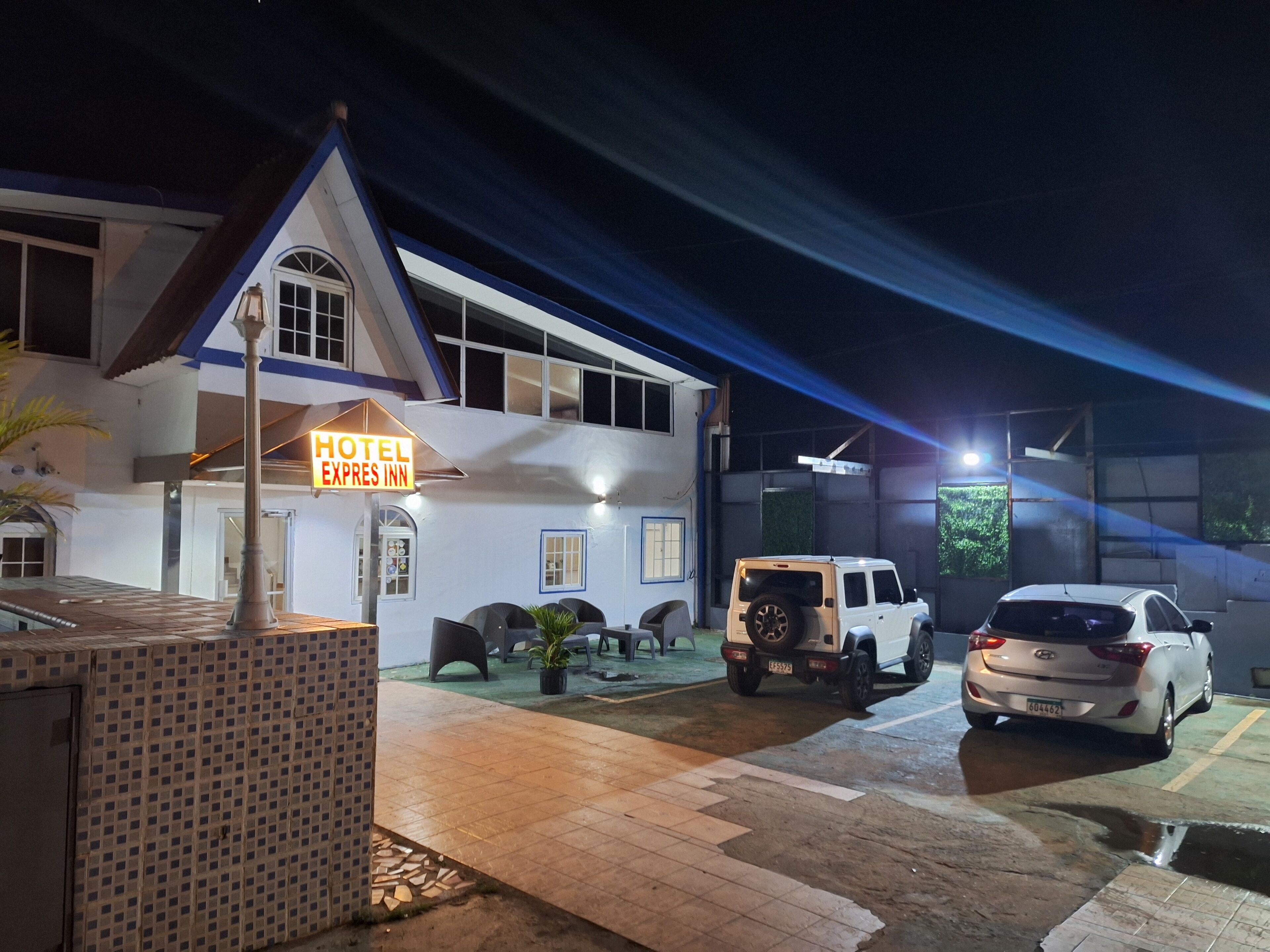 A hotel with a sign reading "Hotel Express Inn," a parking area with two cars, and a building with a tiled roof and a balcony.