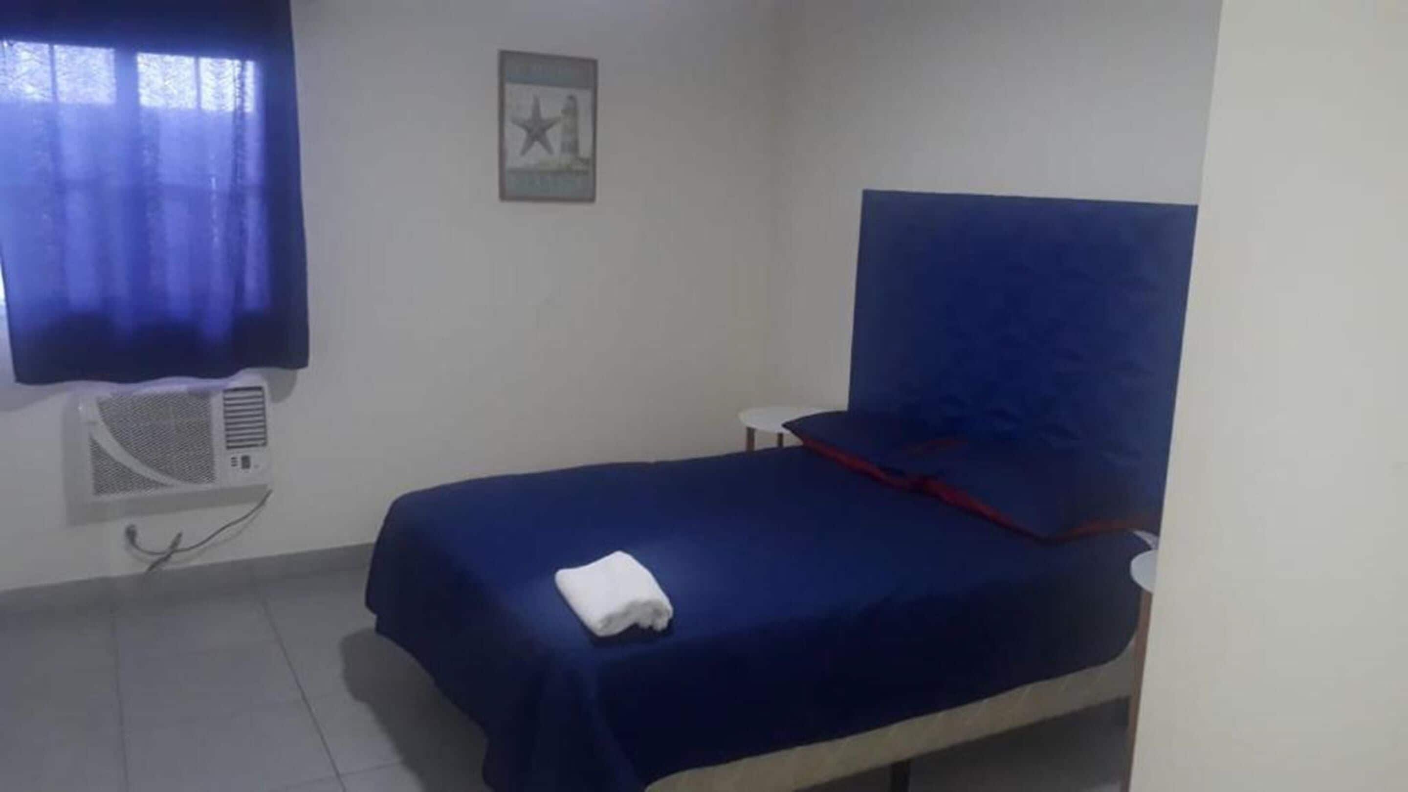 Un lit simple avec un matelas bleu et une couverture blanche, une petite table de chevet, un climatiseur fixé au mur et une fenêtre avec un rideau bleu.