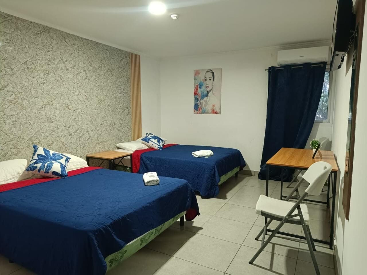Una habitación de hotel con dos camas, una mesita, una silla y un cuadro en la pared.