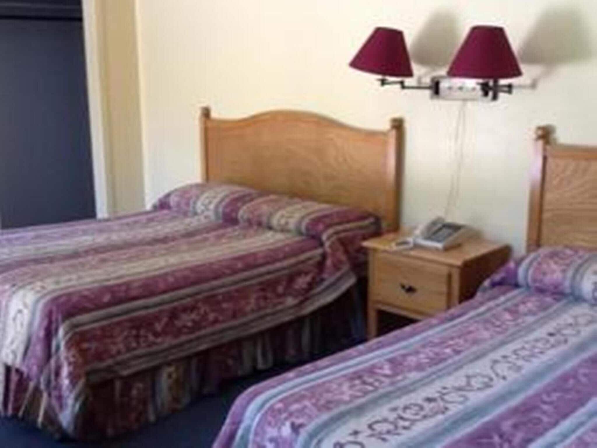 A hotel room with two beds, each with patterned bedding. There is a bedside table with a lamp and a phone.