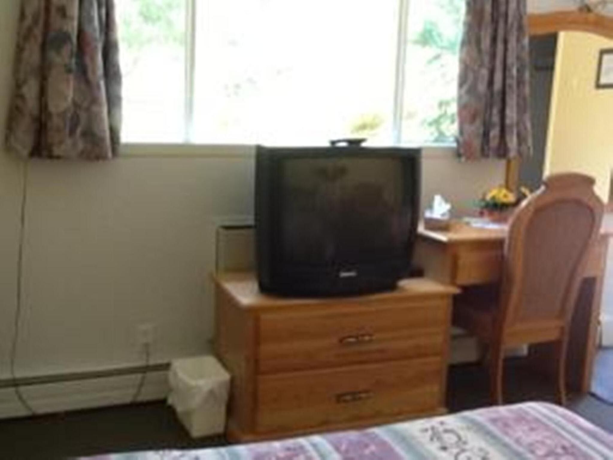 Una habitación con un escritorio de madera, una silla, un televisor sobre un aparador de madera y una ventana con cortinas.