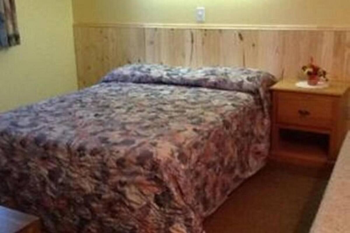 Un lit simple avec une couvre-lit à motifs floraux, dans une pièce aux murs recouverts de lambris en bois et agrémentée d’une table de chevet avec une lampe.