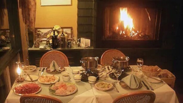 A dining table set for a meal with various dishes and utensils, surrounded by wicker chairs, with a fireplace in the background. - Auberge Mountain View Inn (Mont-Tremblant)