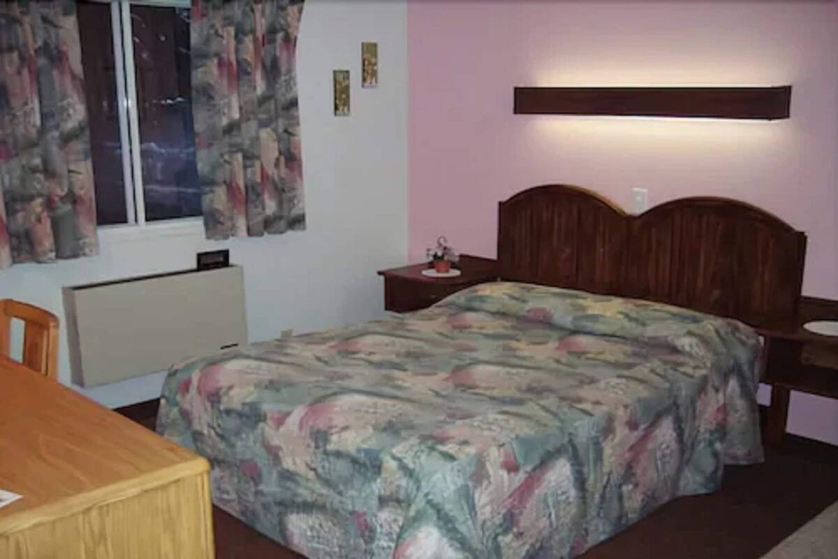 Une chambre à coucher comprenant un lit, une table de chevet, une chaise, une fenêtre avec des rideaux et un radiateur.